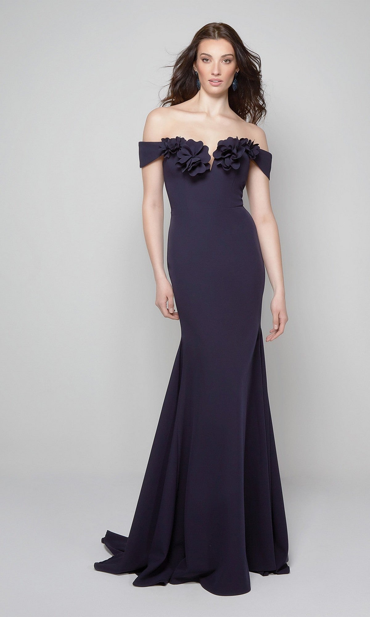 A woman models the Alyce 7062 long formal dress in midnight, featuring an off-the-shoulder neckline with 3D floral accents. She stands against a plain light background. Ideal attire for elegant evening events.